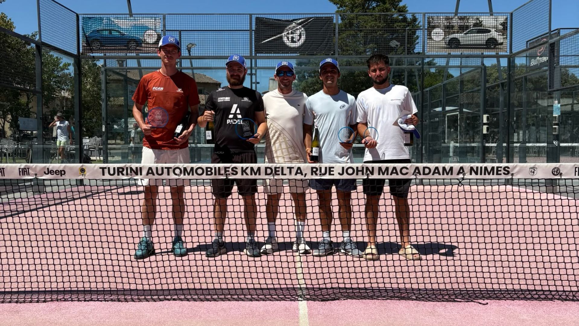 Five men in blue caps stand side by side on a pink outdoor court, holding trophies and bottles after a match.