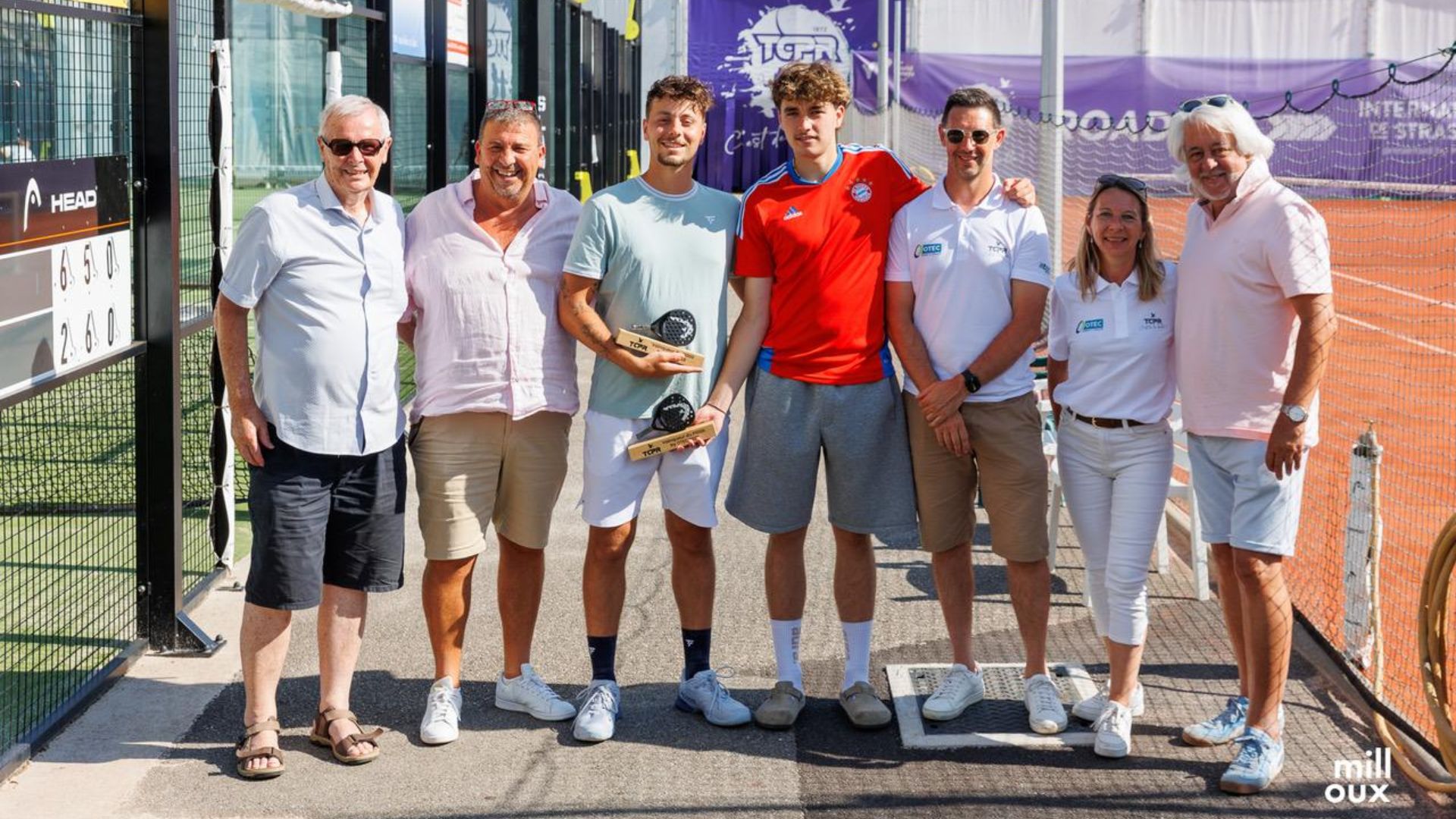 Eight people pose together on a sunny tennis court after a trophy ceremony, two men holding wooden trophies.