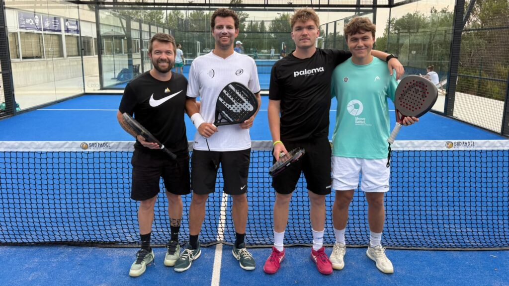 Four men stand together on a blue padel court, holding paddles and smiling at the camera.