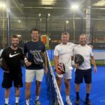 Four men standing side by side on a bright blue indoor padel court, each holding a racket and smiling after a game.