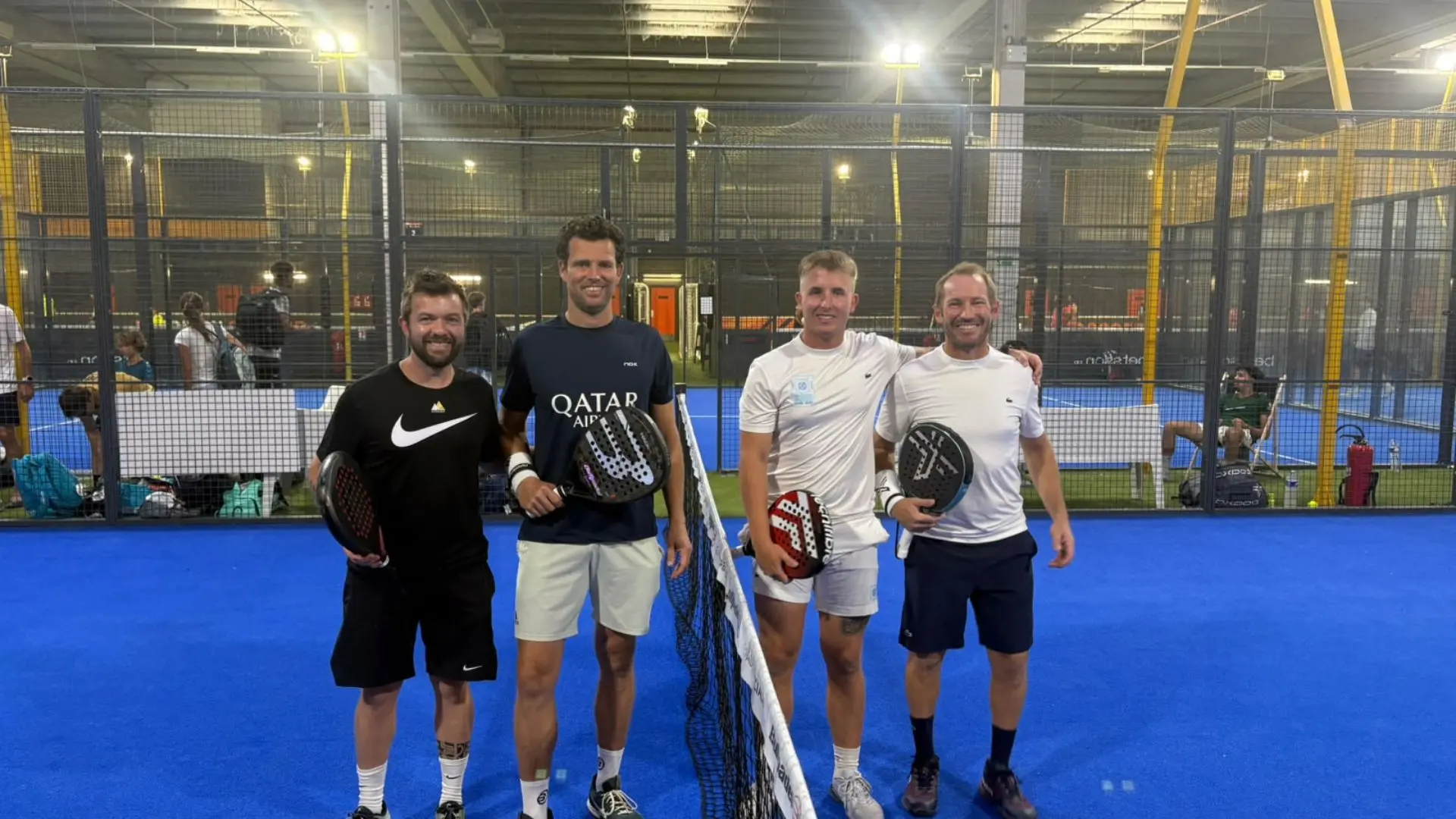 Four men standing side by side on a bright blue indoor padel court, each holding a racket and smiling after a game.