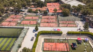 Aerial view of a sports complex with many red clay tennis courts fenced in, surrounded by trees and a parking lot.