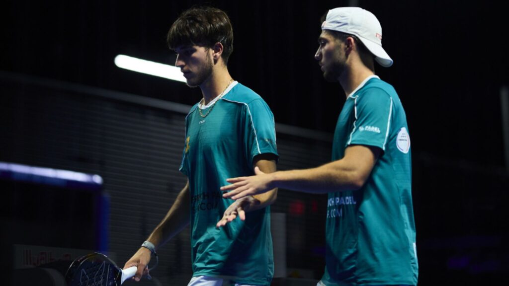 Two male padel players in teal jerseys greet each other at the net, one wearing a white cap backward, indoors.