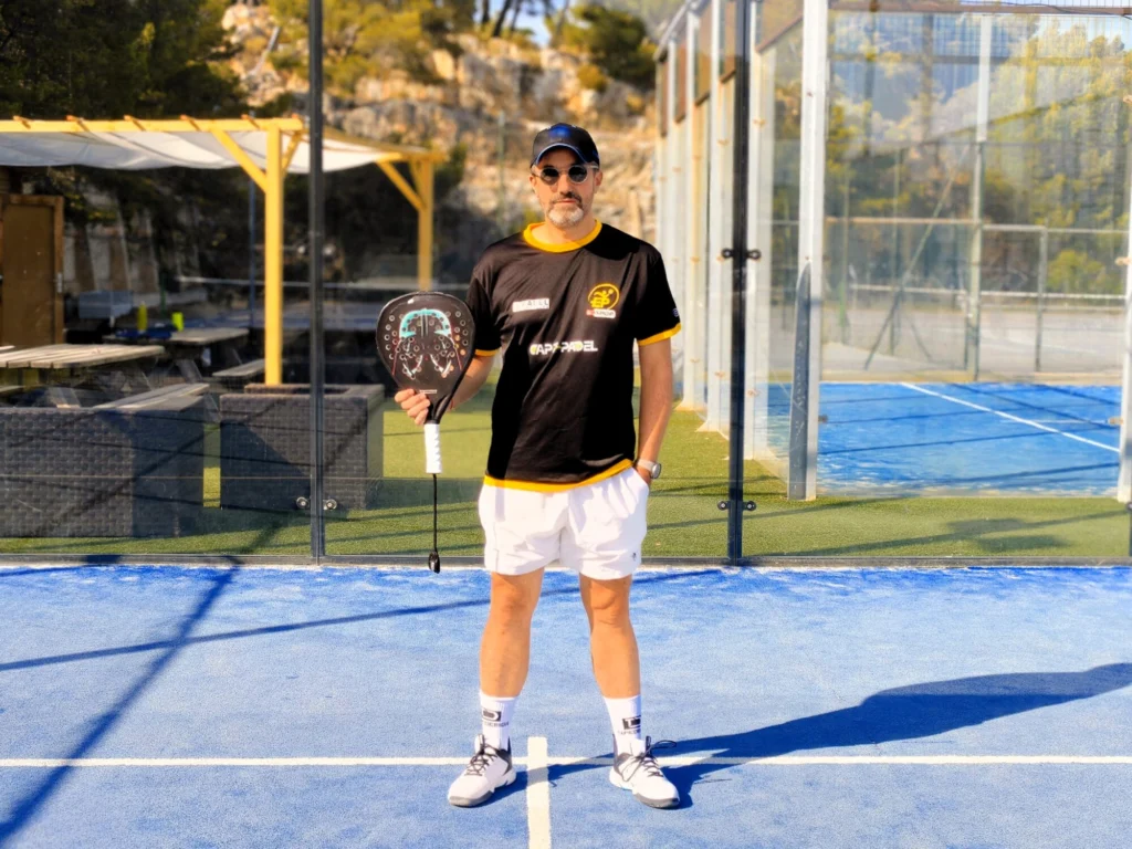 Man standing on a blue padel court, holding a paddle, wearing a black and yellow shirt, cap and sunglasses.