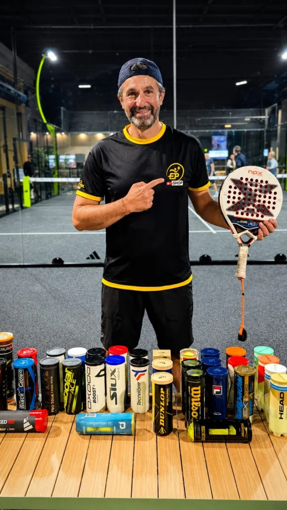 黒と黄色のシャツを着た男性が、スポーツ施設内でパデルラケットを持ち、胸のロゴを指差している。彼の前の木製のテーブルには、飲み物の缶がずらりと並べられている。
