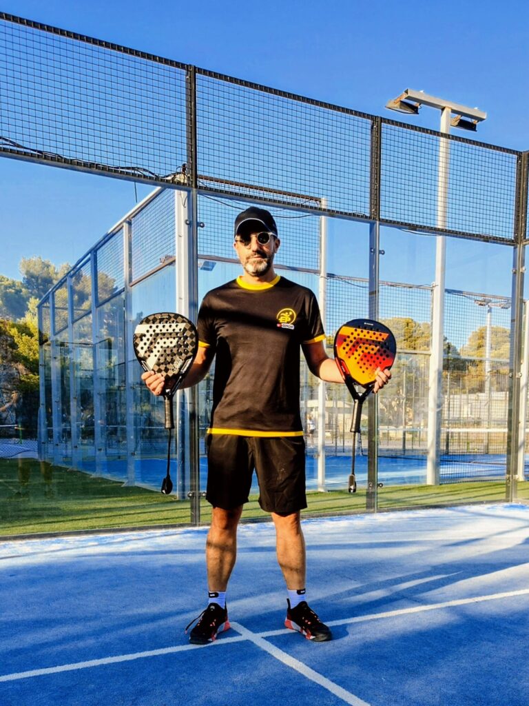 Stef TF1 | Padel Magazine Man on a blue padel court holding two paddles, wearing a black shirt, cap and sunglasses in a sunny setting.