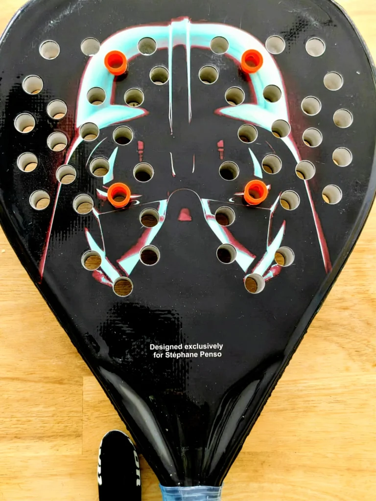 Close-up of a lacrosse stick head with many holes and orange grommets, showing a stylized graphic and the designer's note at the bottom