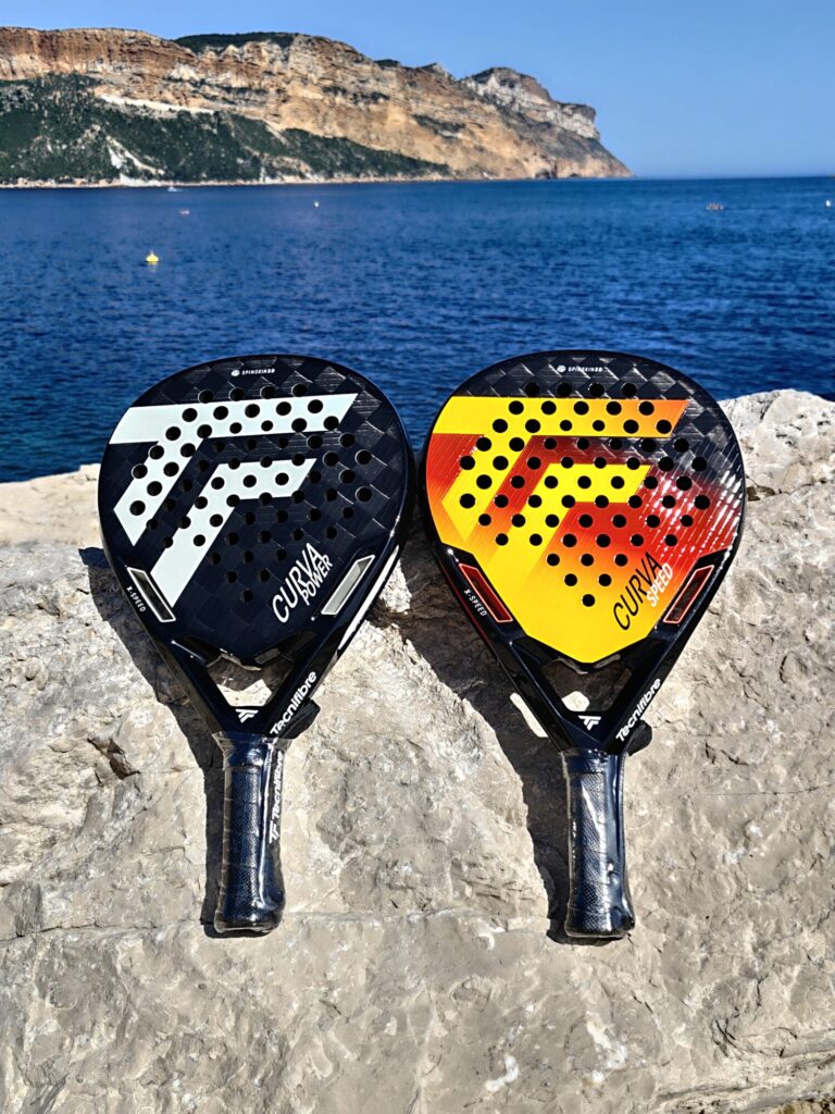 oplus_1075839008 | Padel Magazine Two perforated padel rackets (black/white and yellow/red) lean on a rocky seawall by the blue sea, with cliffs in the background.