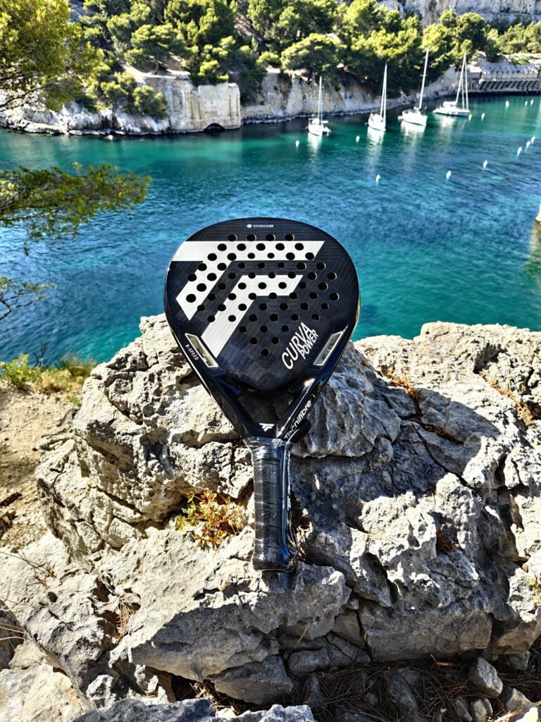 oplus_1075839008 | Padel Magazine Padel racket on a rocky ledge overlooking turquoise water with sailboats anchored along a tree-covered coast.