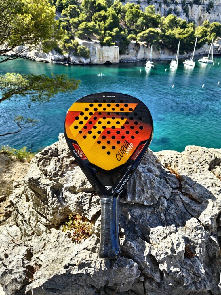 oplus_1210056736 | Padel Magazine Bright yellow and orange paddle resting on jagged rocks overlooking a turquoise cove with sailboats and green hills in the background