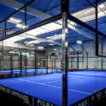 Indoor padel tennis court with blue playing surface, glass walls, and surrounding fencing in a spacious sports facility