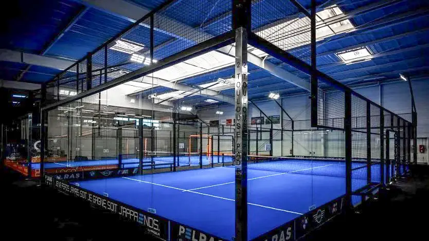 Indoor padel tennis court with blue playing surface, glass walls, and surrounding fencing in a spacious sports facility