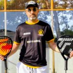 Tecnifibre Curva Dans la lignée dun succès | Padel Magazine Man wearing a black shirt with yellow trim and cap stands on a padel court, holding two padel rackets (orange left, black right). The glass wall and nets are behind him.