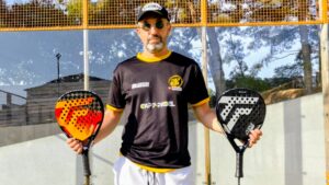 Man wearing a black shirt with yellow trim and cap stands on a padel court, holding two padel rackets (orange left, black right). The glass wall and nets are behind him.