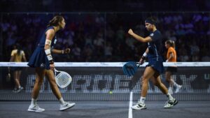 Two female players in blue outfits celebrate mid-match on a padded court, holding perforated paddles near the net as spectators watch from the stands.