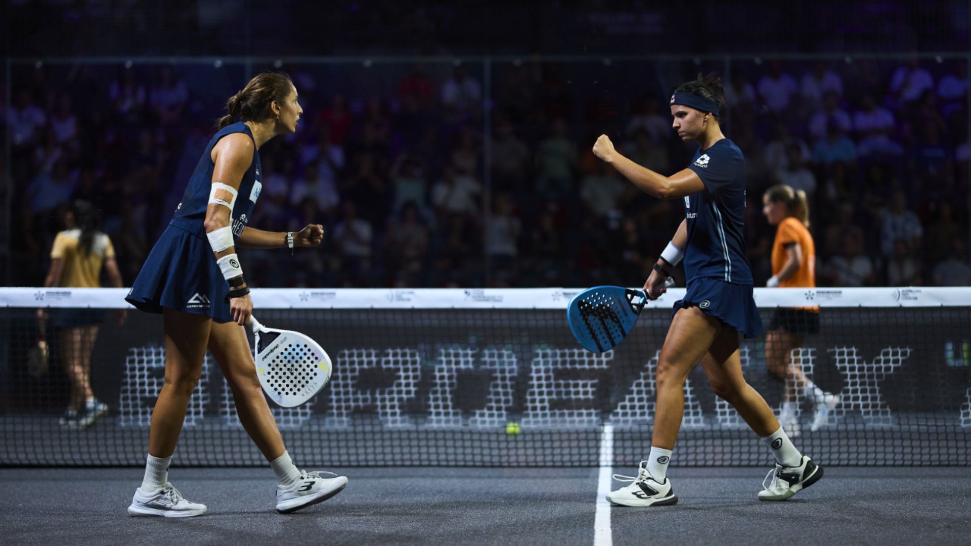Two female players in blue outfits celebrate mid-match on a padded court, holding perforated paddles near the net as spectators watch from the stands.