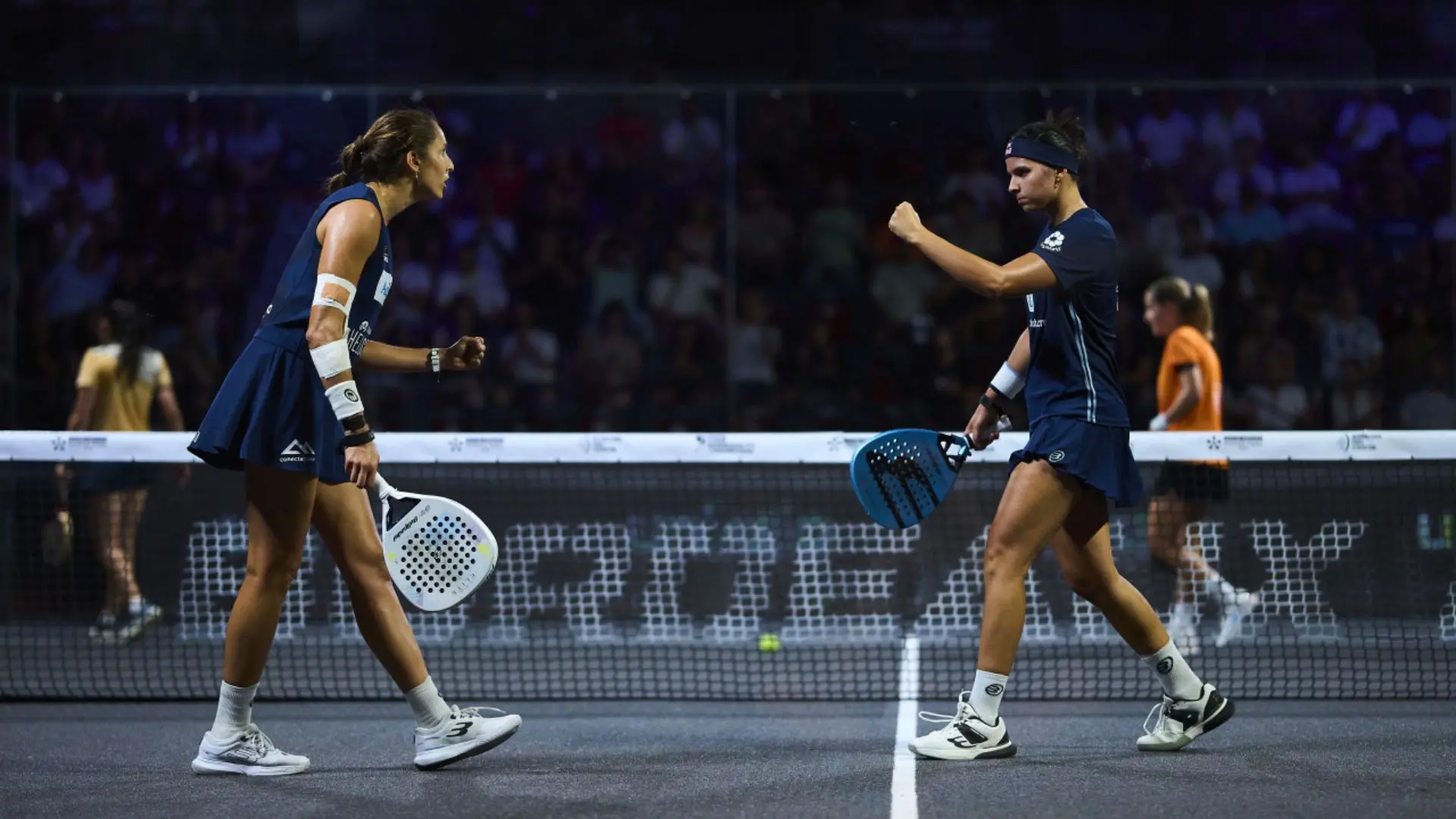 Two female players in blue outfits celebrate mid-match on a padded court, holding perforated paddles near the net as spectators watch from the stands.