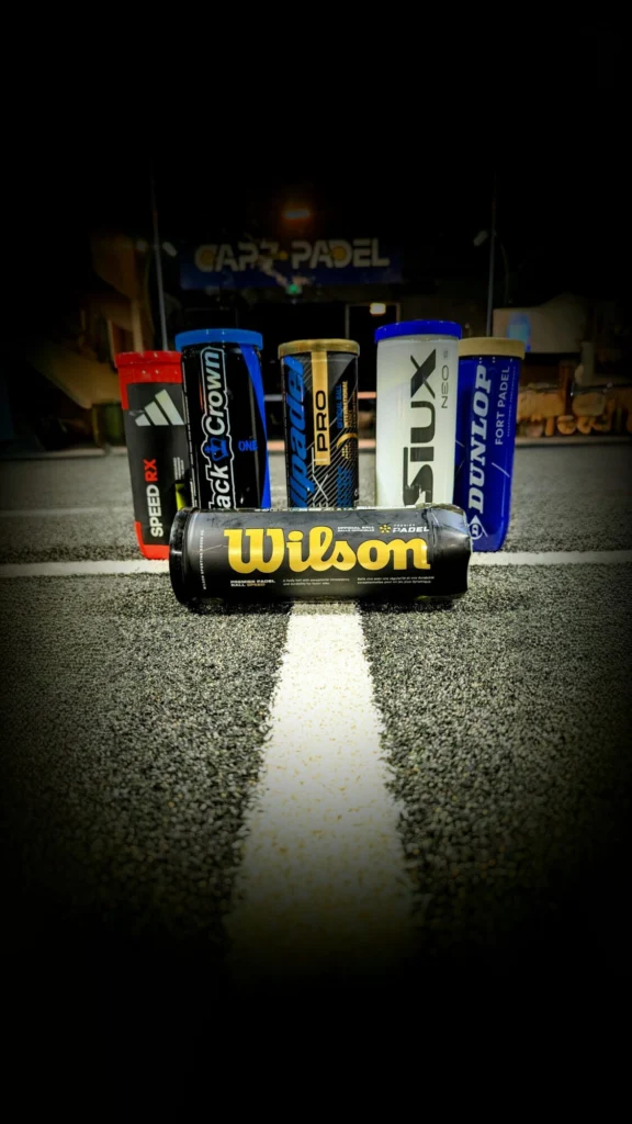 Row of Wilson and other padel ball cans lined up on a padel court at night, foreground white line on the dark surface.