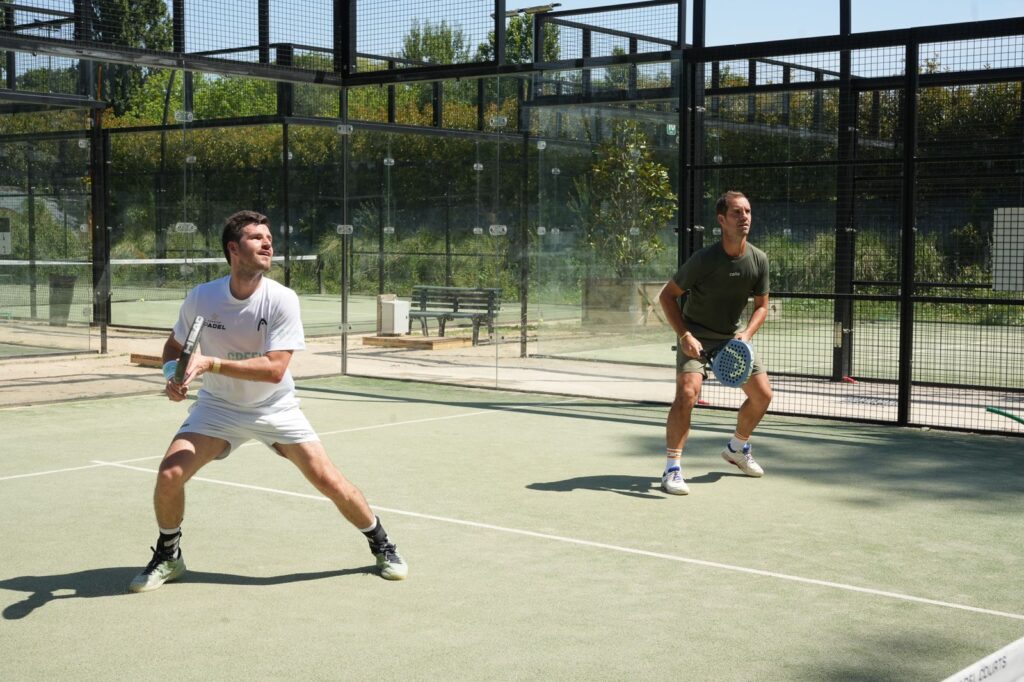 Two men playing padel on an outdoor enclosed court; the white-clad player is preparing a shot while the other watches from the back left.