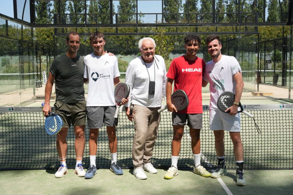 Five men stand shoulder to shoulder on a padel court, each holding a padel paddle and smiling at the camera.