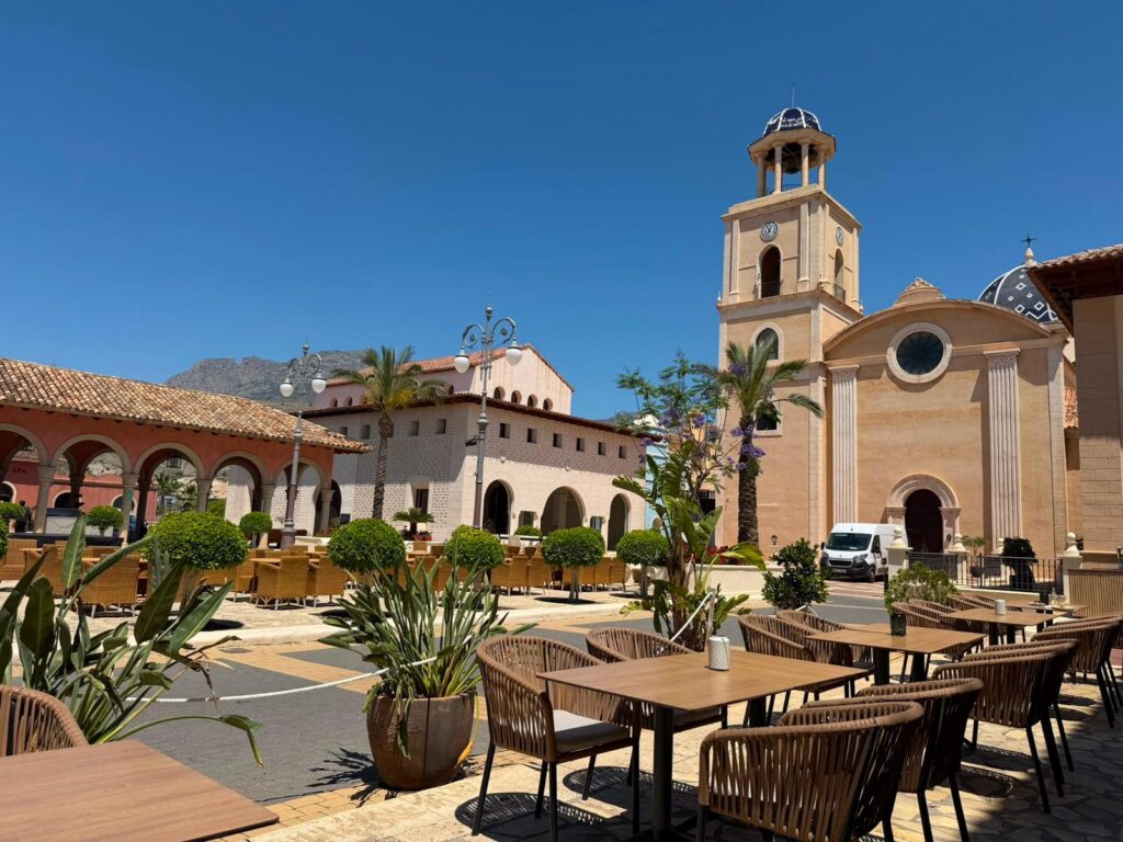 Benidorm, la nouvelle destination espagnole qui séduit les passionnés de padel | Padel Magazine Outdoor courtyard with a beige church and bell tower, arched colonnade, and wicker tables under a clear blue sky.
