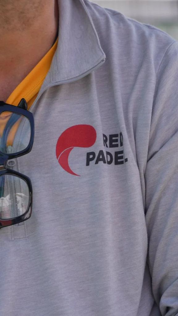 Close-up de uma pessoa vestindo uma camisa polo cinza-clara com o logotipo vermelho da Red Paddle e uma camisa amarela por baixo; óculos de sol pendurados na gola.