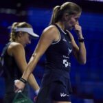 Two female tennis players in dark outfits walk on a blue-lit indoor court; foreground player bites her nails and wears a ponytail.