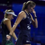 collombon sharifova malaga p1 1 | Padel Magazine Two female tennis players in dark outfits walk on a blue-lit indoor court; foreground player bites her nails and wears a ponytail.