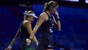 Two female tennis players in dark outfits walk on a blue-lit indoor court; foreground player bites her nails and wears a ponytail.