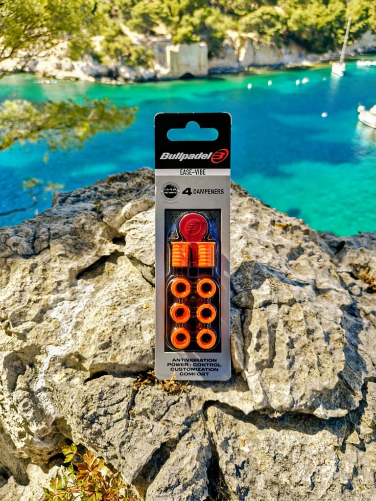 Pack of Bullpadel Ease-Vibe dampeners in orange, displayed on a rocky coastline with turquoise water and boats in the background.