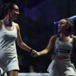 Two female padel players in white outfits shake hands on a court after a match, smiling at each other.