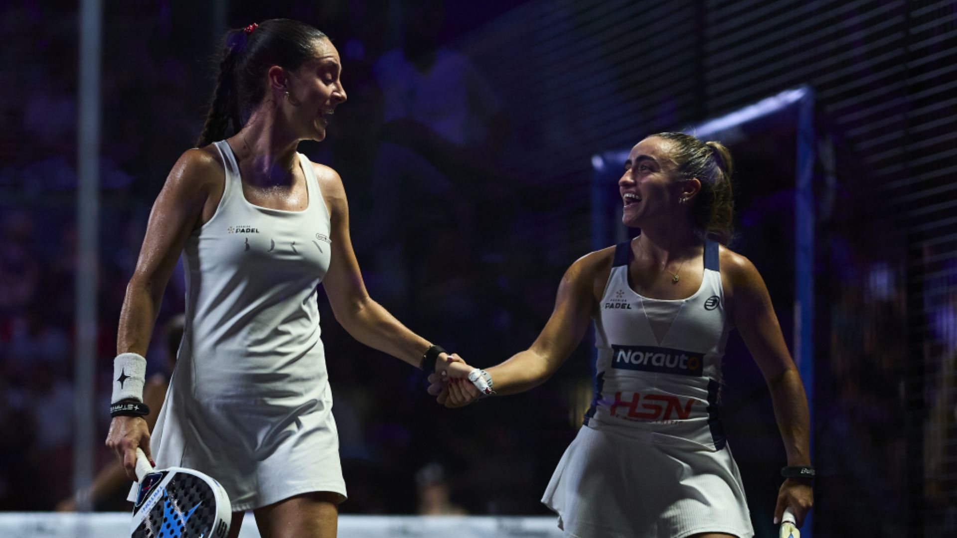 Two female padel players in white outfits shake hands on a court after a match, smiling at each other.