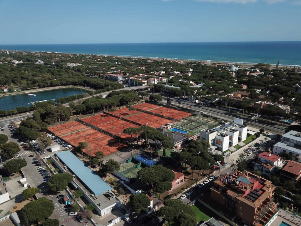 Aerial view of a coastal town with orange clay tennis courts in the foreground, green trees, and the blue sea on the horizon.