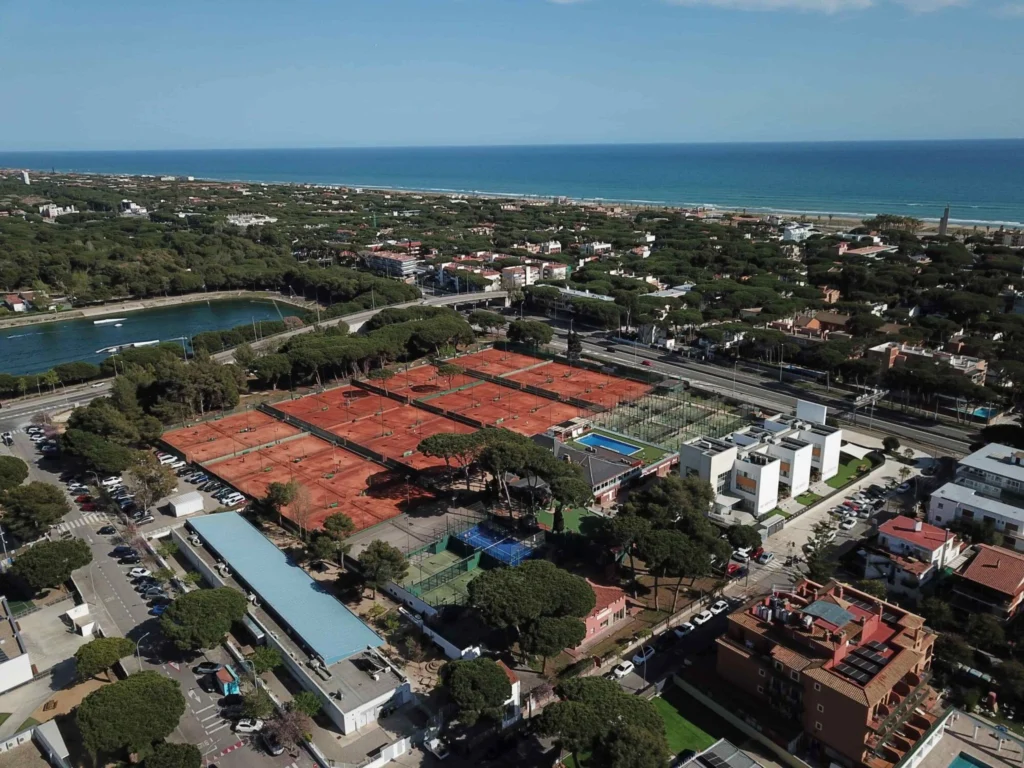 image | Padel Magazine Aerial view of a coastal town with orange clay tennis courts in the foreground, green trees, and the blue sea on the horizon.