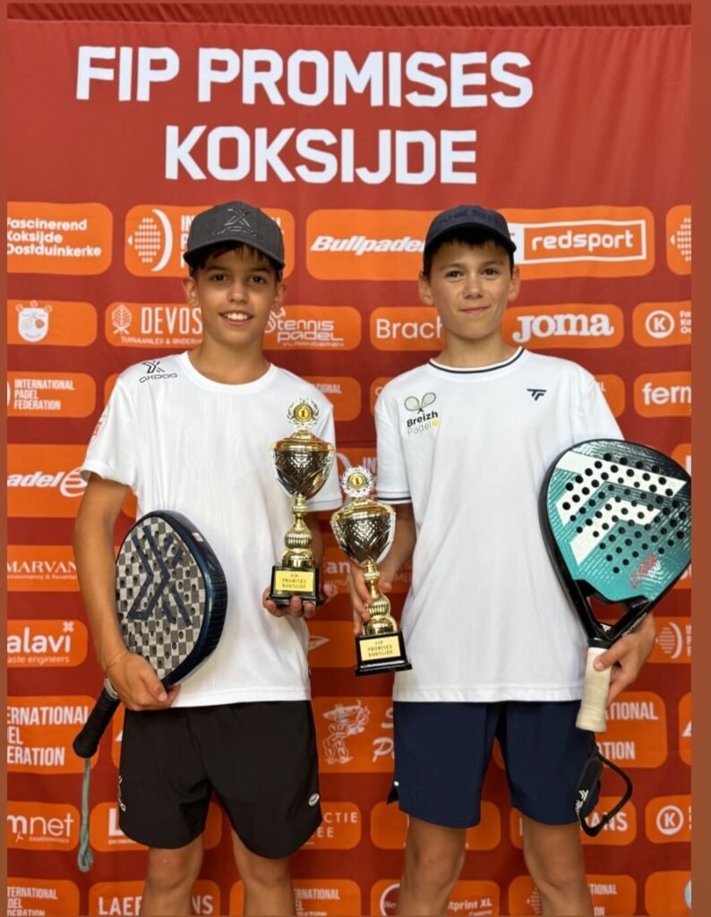 Two boys in white shirts hold two trophies and paddles, posing for a photo at a paddle tennis event with an orange sponsor backdrop.