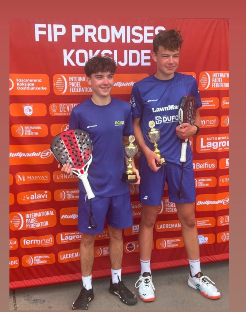 Two young padel players in blue uniforms, smiling, holding trophies and paddles in front of a red sponsor backdrop.