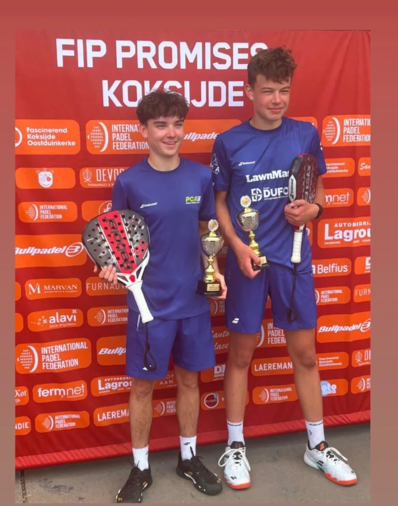 Two young padel players in blue uniforms, smiling, holding trophies and paddles in front of a red sponsor backdrop.