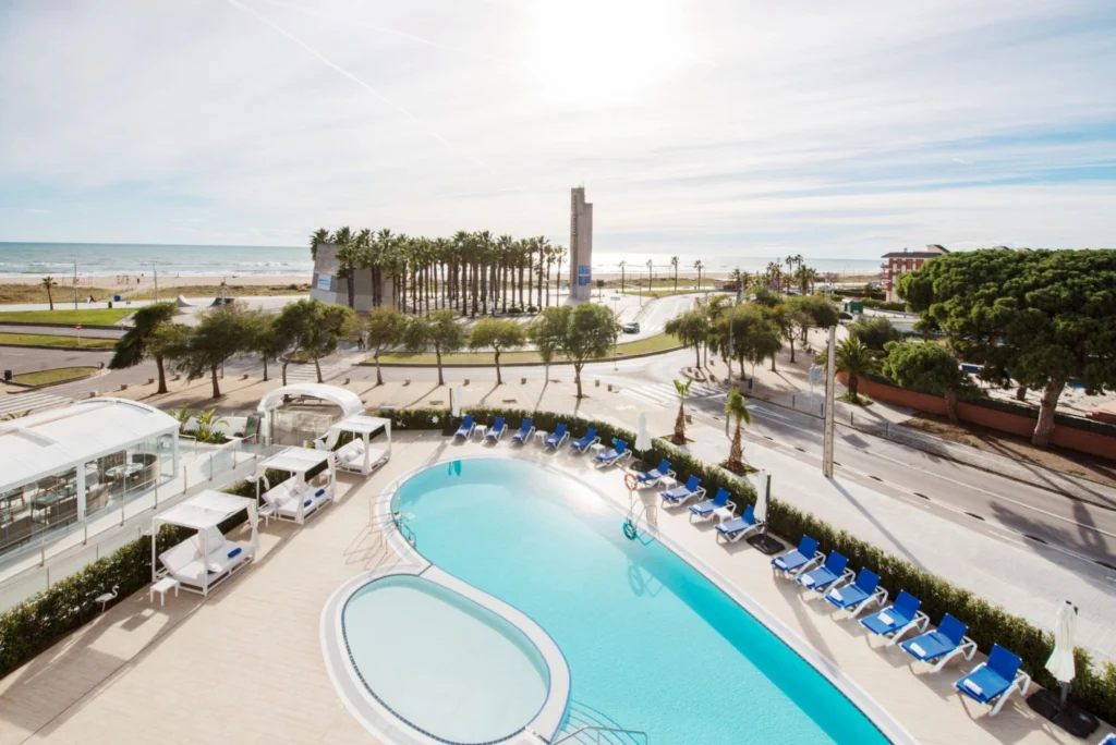image | Padel Magazine Pool deck at a seaside hotel with blue lounge chairs, a curved main pool, a circular plunge pool, palm trees, and the beach and ocean in the background.