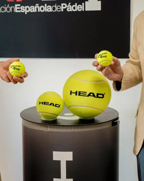 bild | Padel Magazine Tre gula HEAD padelbollar visas på en rund svart piedestal, med två personer som placerar mindre bollar ovanpå, och en bakgrund som visar en del av en logotyp.