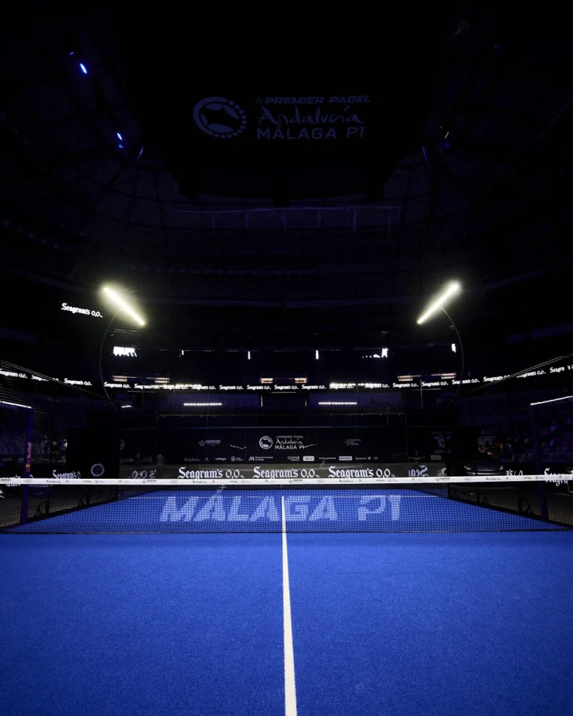 Blue padel court in a dark arena with a net across the center and MALAGA P1 branding on the surface.