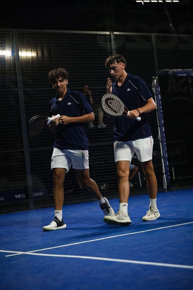 Two male padel players walk on a blue court, holding paddles and towels after a match indoors.