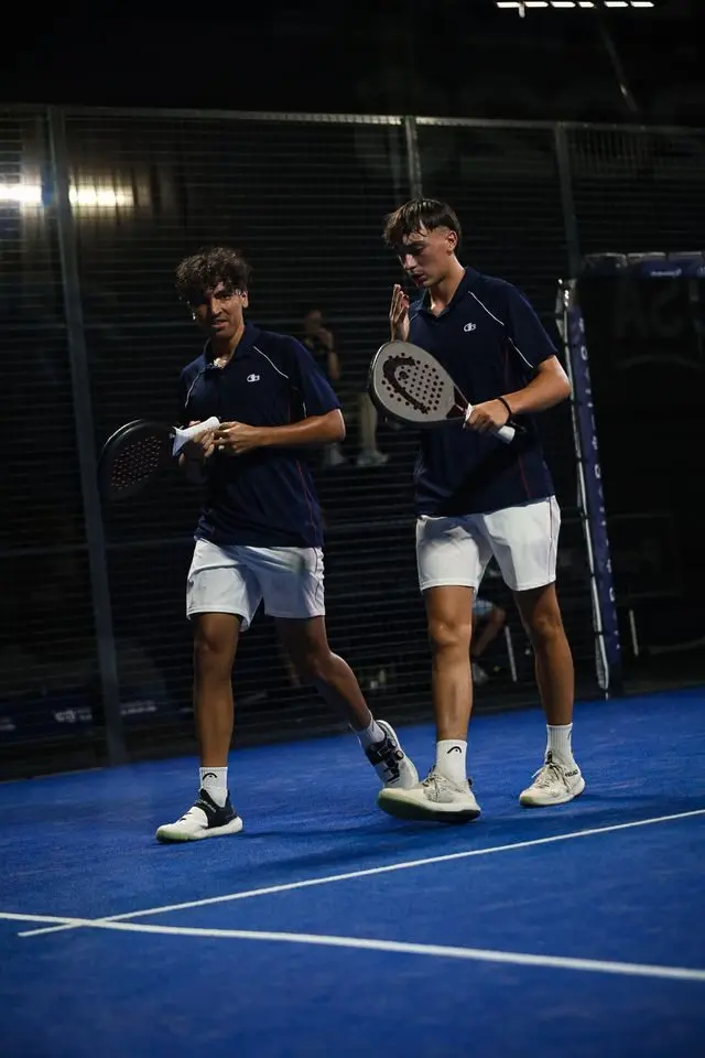 image | Padel Magazine Two male padel players walk on a blue court, holding paddles and towels after a match indoors.