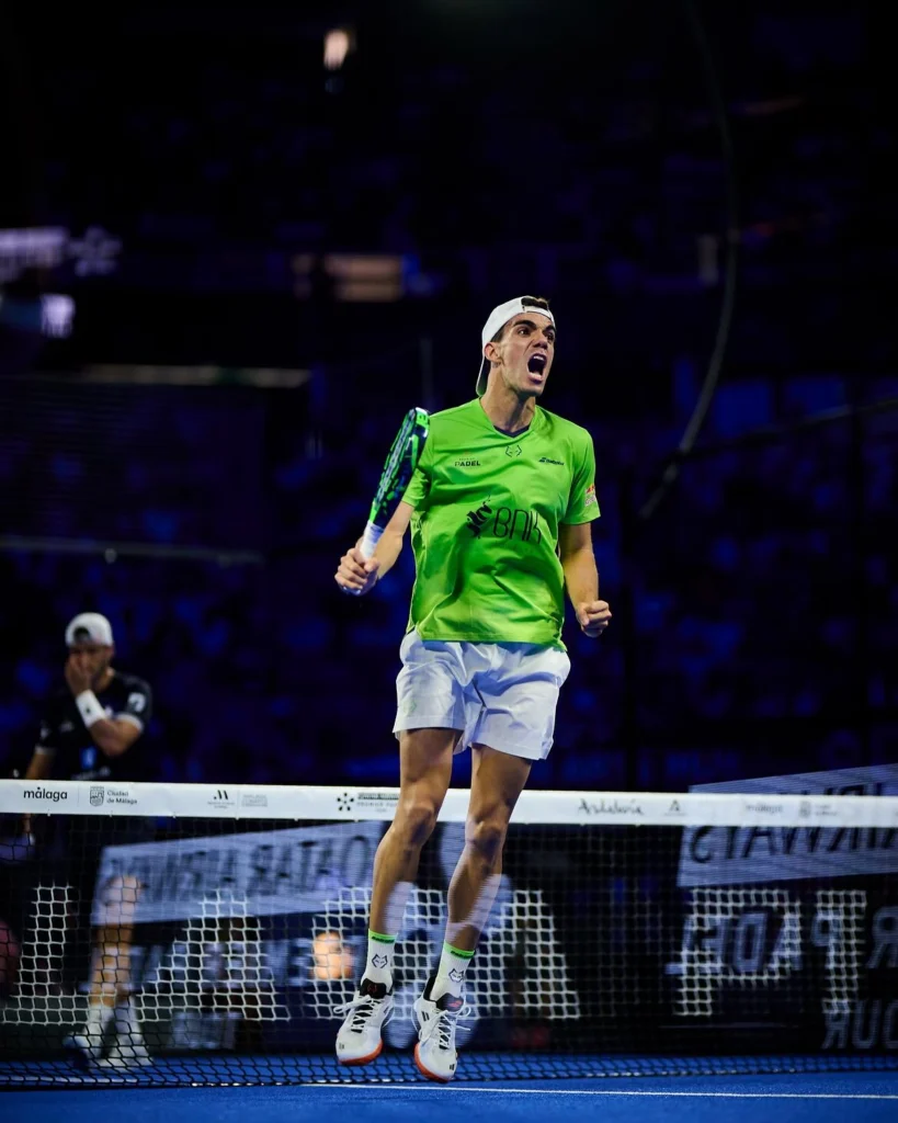immagine | Padel Magazine Un giocatore di padel con indosso una maglietta verde salta a mezz'aria con la racchetta, festeggiando su un campo blu vicino alla rete.