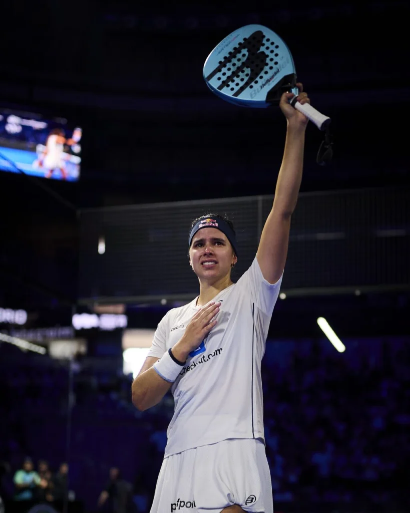 image | Padel Magazine Female padel player raises a blue paddle overhead in a victory pose during a match.
