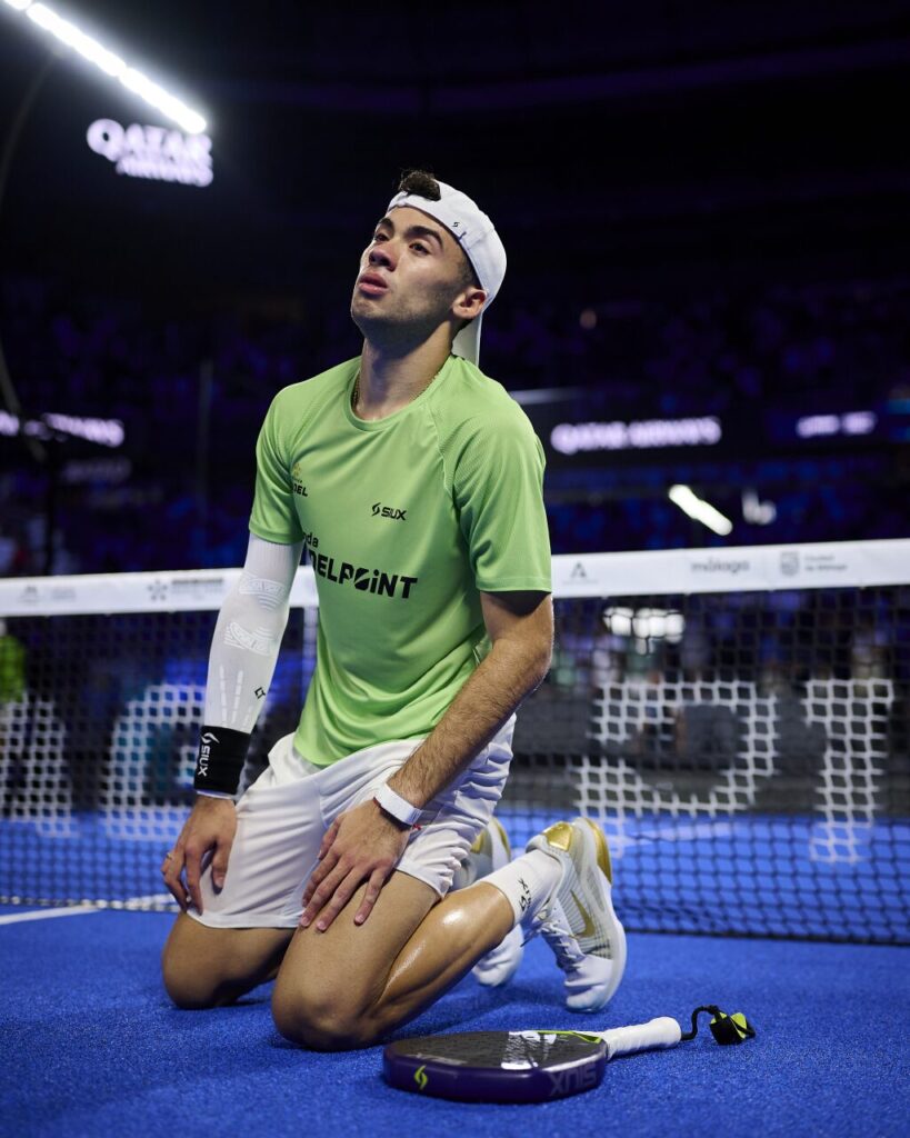 Male athlete kneeling on a blue court with a paddle beside him, wearing a green shirt and white cap.