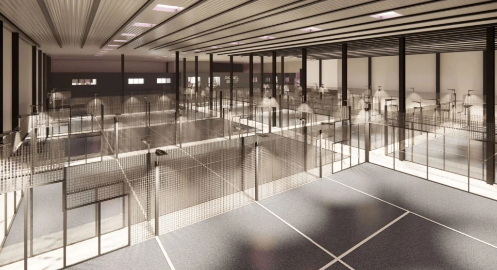 picture | Padel Magazine A spacious indoor sports hall with multiple court lanes separated by metal mesh partitions, tall black columns, and bright overhead lighting viewed from an upper level central walkway.
