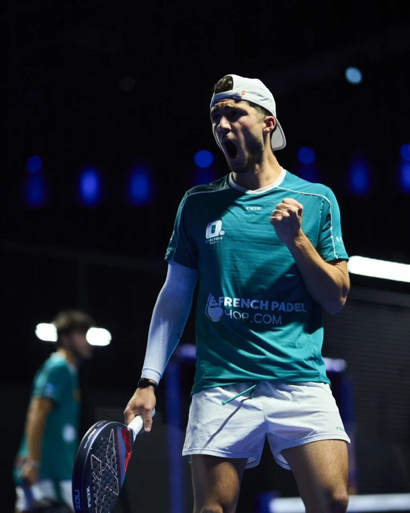 image | Padel Magazine Padel player in teal jersey and white cap, fist pumped in celebration on an indoor court with blue lighting.