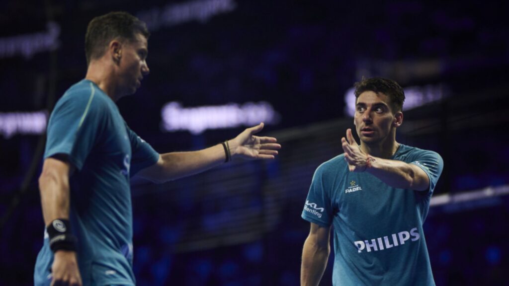 Two male padel players in blue shirts high-fiving on a dimly lit court.