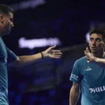 Two male padel players in blue shirts high-fiving on a dimly lit court.