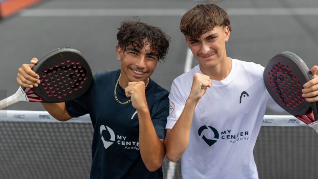 Two young men on a padel court pose with fists up, holding rackets, wearing My Center Academy shirts and smiling at the camera.